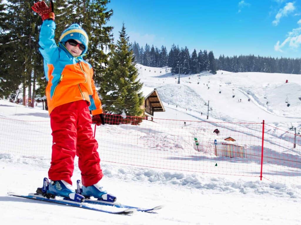 Enfant heureux en tenue de ski colorée, en train de skier sur une piste ensoleillée pendant ses vacances d’hiver en famille.