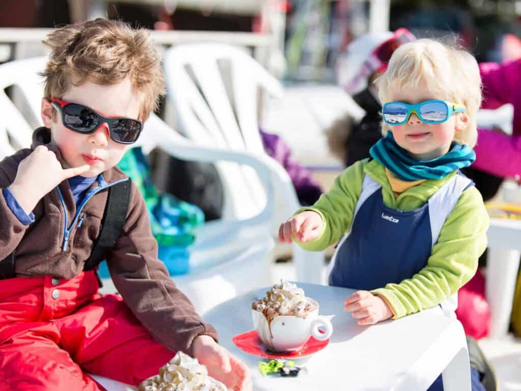 Deux enfants ravis en tenue de ski dégustant un chocolat chaud dans un café pendant leurs vacances d'hiver en famille