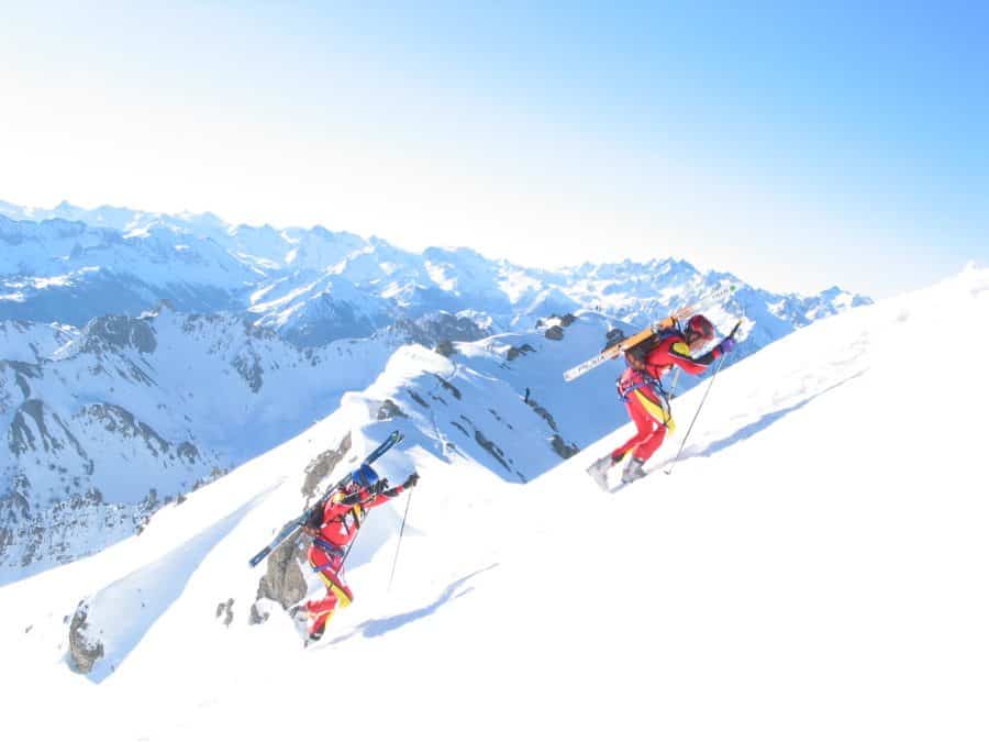 Two men walk through the snow carrying their skis on their backs