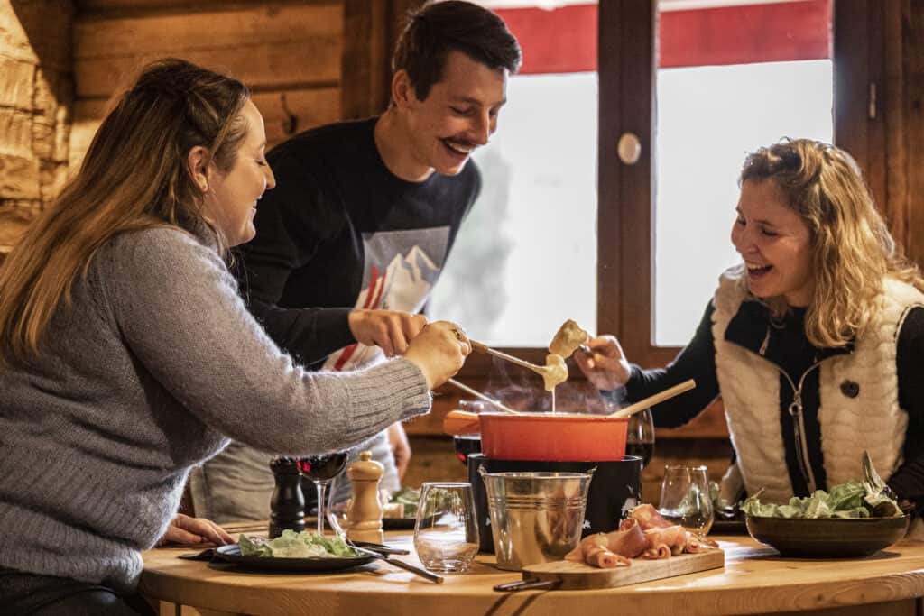 Un homme et deux femmes autour d'une table, souriants en train de déguster une fondue. 