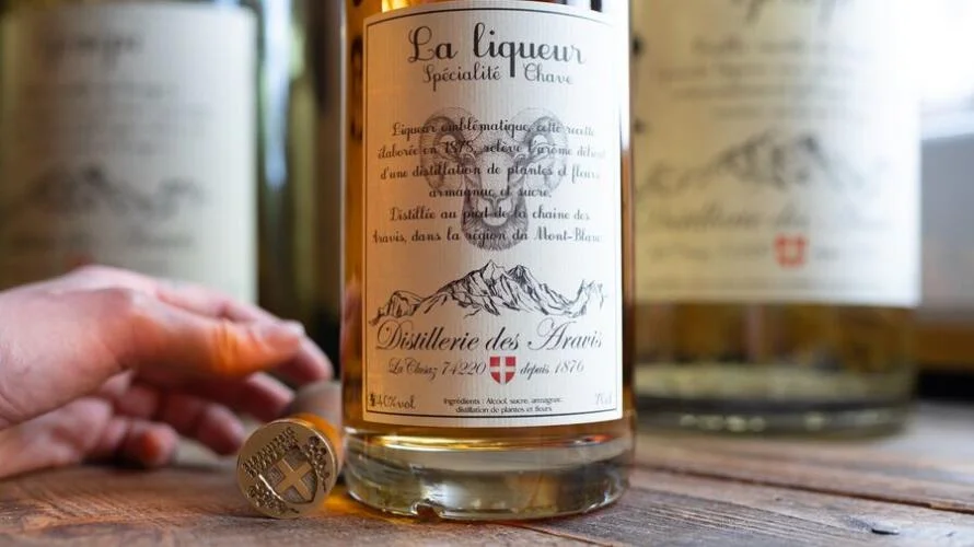 A bottle of liqueur from the Distillerie des Aravis and a hand holding the seal of the distillery.