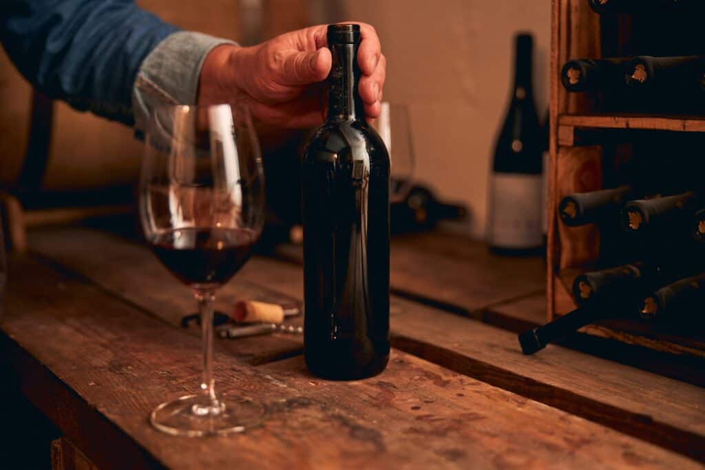 Un homme manipule une bouteille et un verre de vin rouge présentés sur une table en bois.