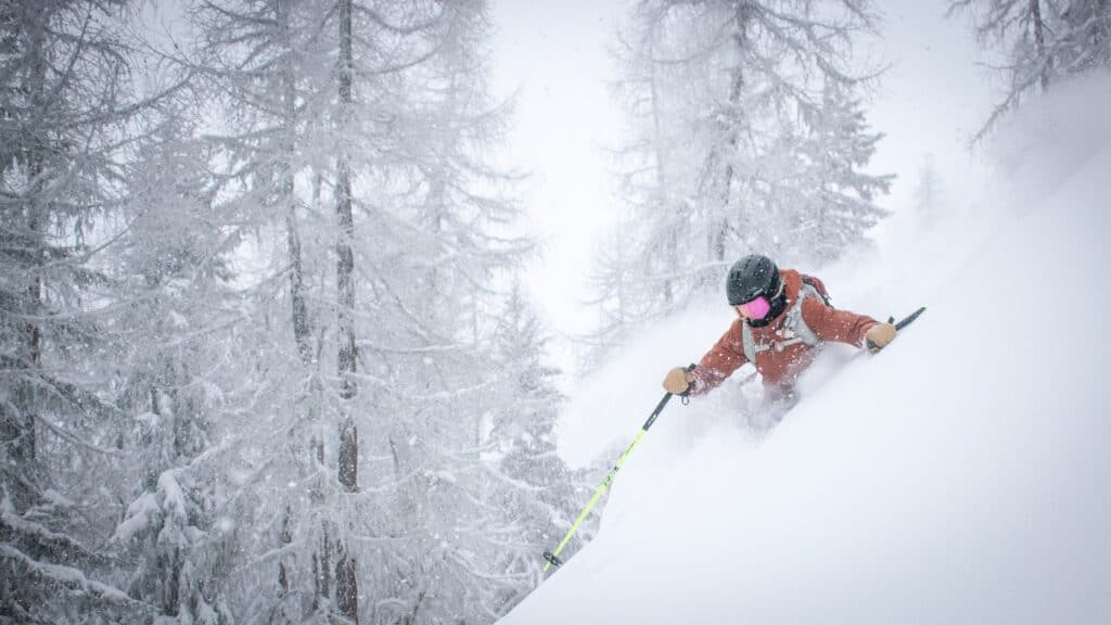 A man in an orange ski jacket skis down an off piste run with lots of powder