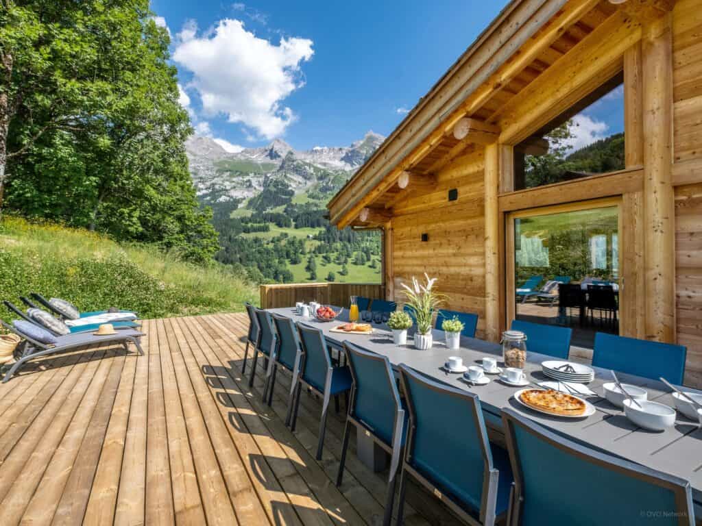 Grande table à manger sur la terrasse d'un chalet boisé, avec montagnes en arrière-plan.